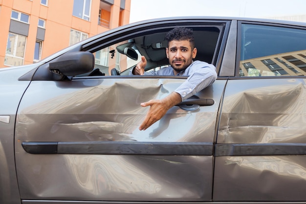 Ritratto di un bell'uomo barbuto confuso che indossa una camicia blu seduto al volante di un'auto ammaccata con un'espressione facciale perplessa, che mostra ammaccature e graffi della portiera dell'automobile. Tiro all'aperto.