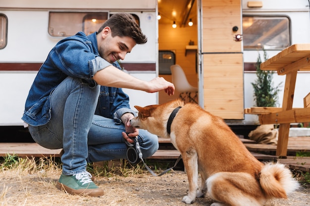 Ritratto di un bell'uomo con la barba lunga che indossa abiti in denim accarezzando un cane rosso mentre si accovaccia vicino a casa su ruote all'aperto