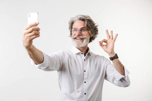 Ritratto di un bell'uomo d'affari maturo felice vestito in abiti formali in piedi isolato su un muro bianco, facendo un selfie, mostrando ok