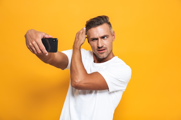 Ritratto di un bell'uomo di 30 anni in maglietta bianca che gesticola e si fa selfie sul cellulare isolato su giallo