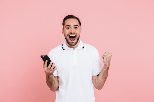 Ritratto di un bell'uomo felice in piedi isolato sopra il rosa, con in mano il cellulare, celebrando il successo