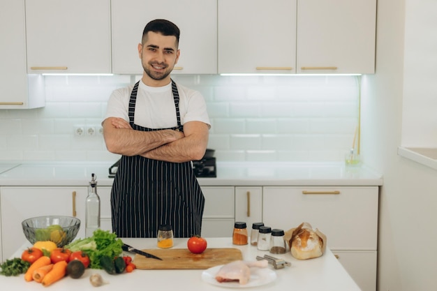 Ritratto di un bell'uomo in cucina Un uomo vestito con un grembiule nero Guarda la telecamera Sul tavolo della cucina stanno cucinando prodotti Pollo Verdure Spezie