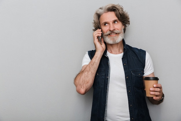 Ritratto di un bell'uomo maturo ed elegante sorridente che indossa un giubbotto in piedi isolato su uno sfondo grigio, parlando al telefono cellulare mentre beve caffè da asporto