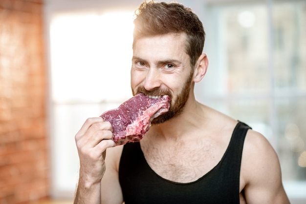 Ritratto di un bell'uomo sportivo con una maglietta nera che morde una bistecca di carne cruda al chiuso