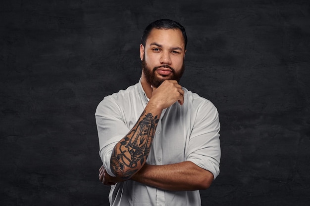 Ritratto di un bell'uomo tatuato afroamericano con capelli e barba alla moda in una camicia bianca, tenendo la mano sul mento. Isolato su uno sfondo scuro.