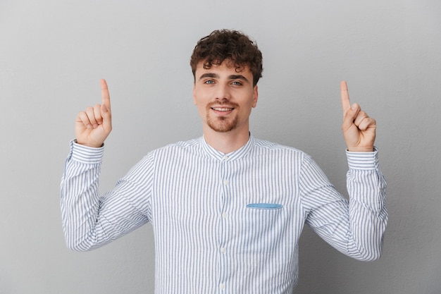 Ritratto di un bell'uomo vestito con una camicia che sorride e punta le dita verso l'alto al copyspace isolato su un muro grigio