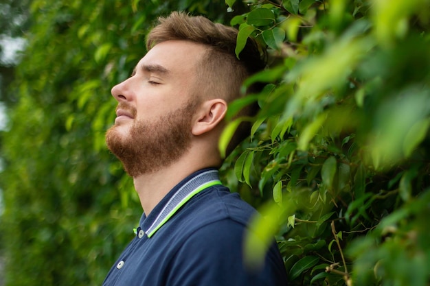 Ritratto di un bellissimo giovane uomo barbuto con la barba in piedi su un muro di edera o nel verde sorridente contro il ragazzo delle piante che si rilassa all'aperto nella soleggiata giornata estiva Ama la natura eco vita