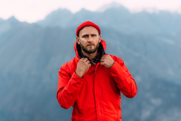 Ritratto di un brutale uomo barbuto con una giacca rossa e un cappello tra le montagne. Ritratto maschile sullo sfondo di un paesaggio di montagna. Un viaggiatore sullo sfondo delle montagne.