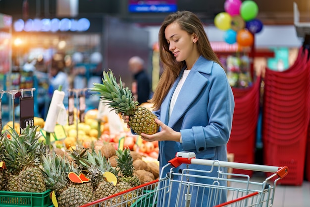 Ritratto di un compratore attraente felice sorridente della donna con il carrello nella drogheria durante la scelta e l'acquisto di ananas fresco al reparto frutta