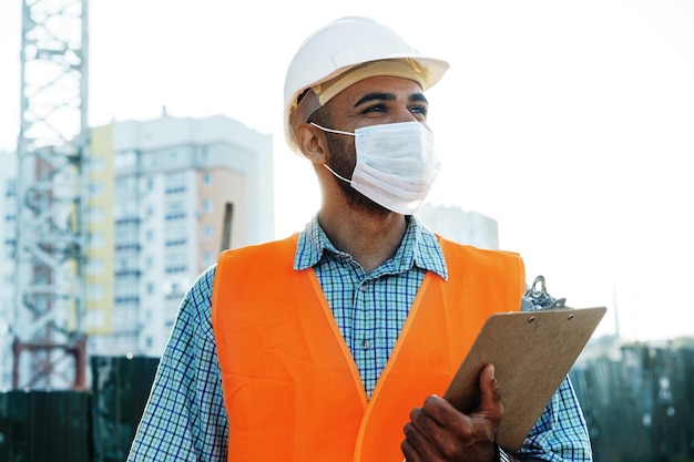 Ritratto di un costruttore di uomini di razza mista in abiti da lavoro e elmetto protettivo che indossa una maschera medica da vicino