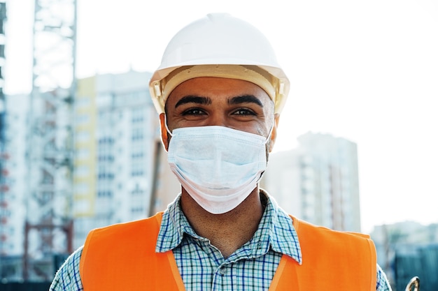 Ritratto di un costruttore di uomini di razza mista in abiti da lavoro e elmetto protettivo che indossa una maschera medica da vicino