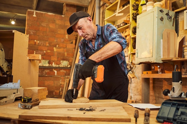 Ritratto di un falegname anziano con strumento di perforazione che lavora nell'officina di falegnameria per realizzare un mobile