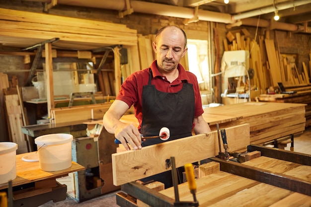 Ritratto di un falegname anziano in uniforme che incolla barre di legno con pressioni manuali presso la produzione di falegnameria