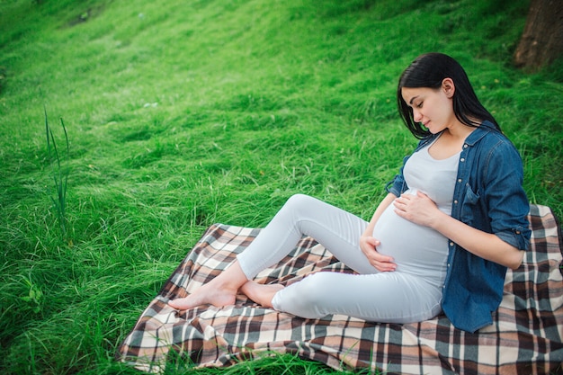 Ritratto di un felice capelli neri e orgogliosa donna incinta in una città nel parco. Foto del modello femminile che tocca la sua pancia con le mani. Il modello femminile è seduto sull'erba.