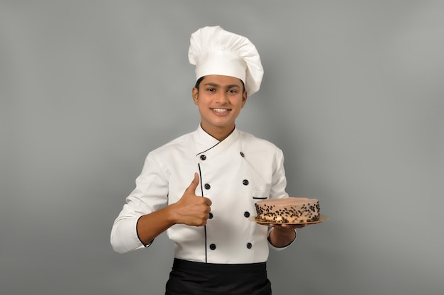 Ritratto di un felice chef maschio vestito in uniforme che tiene piatto con torta al cioccolato con mostra pollice in alto