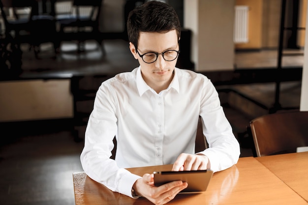 Ritratto di un fiducioso giovane manager seduto in una caffetteria che lavora su un tablet vestito con gli occhiali da vista.