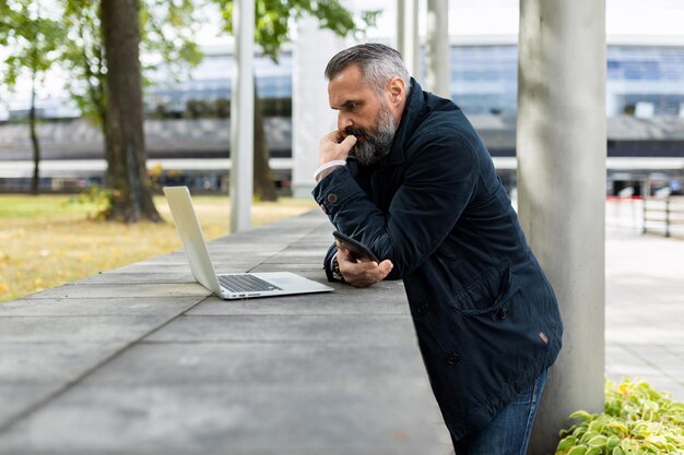 Ritratto di un finanziere maschio adulto maturo con barba e capelli grigi lavora con un computer portatile in città