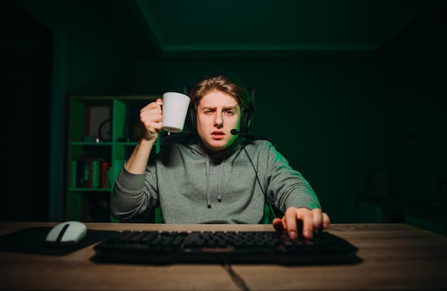 Ritratto di un giocatore stanco in un auricolare con una tazza in mano che gioca ai giochi online