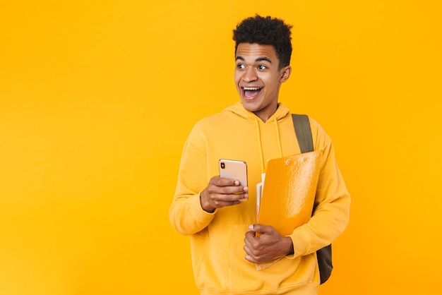 Ritratto di un giovane adolescente felice in piedi isolato su un muro giallo, che trasporta zaino e libri di testo, utilizzando il telefono cellulare