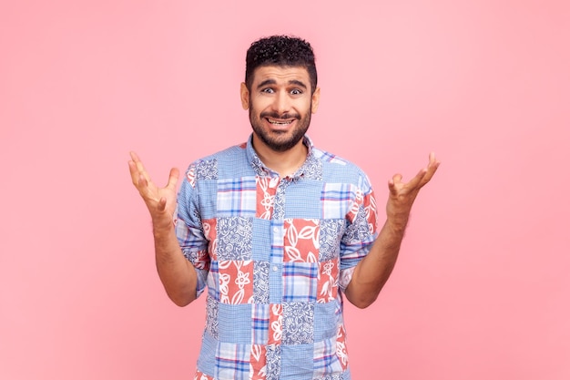 Ritratto di un giovane adulto confuso o scioccato con la barba in camicia blu casual in piedi con le braccia alzate e guardando la fotocamera chiedendo. Studio indoor girato isolato su sfondo rosa.