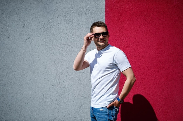 Ritratto di un giovane alla moda, un uomo vestito con una maglietta bianca vuota in piedi su uno sfondo grigio e rosso della parete. Stile urbano di vestiti, immagine alla moda moderna. Moda maschile