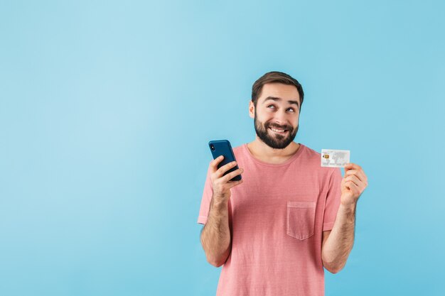 Ritratto di un giovane allegro eccitato uomo barbuto che indossa una t-shirt in piedi isolato su un muro blu, utilizzando il telefono cellulare, mostrando la carta di credito, facendo shopping