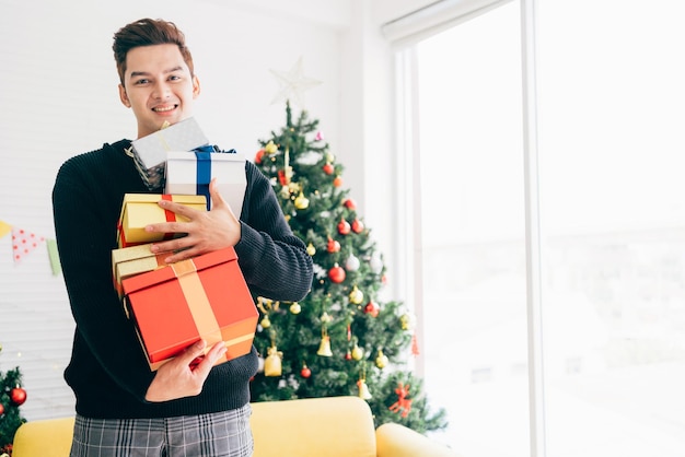 Ritratto di un giovane asiatico eccitato e sorpreso che tiene molti regali che si accatastano nelle sue mani nel soggiorno con albero di Natale decorato sullo sfondo