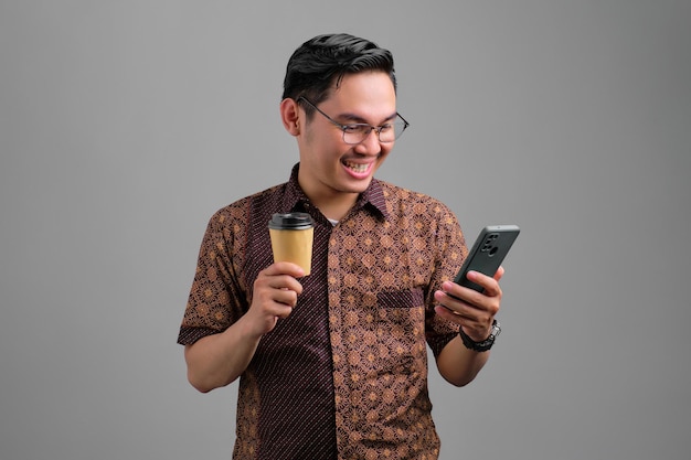 Ritratto di un giovane asiatico sorridente che indossa una camicia batik utilizzando lo smartphone che tiene una tazza di caffè di carta isolata su sfondo grigio