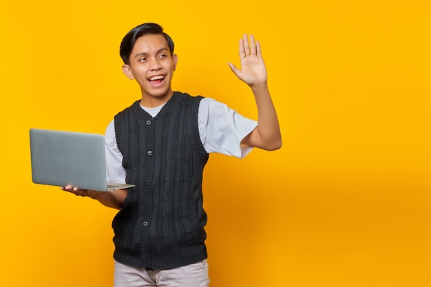 Ritratto di un giovane asiatico sorridente che tiene in mano un laptop e fa un gesto con la mano d'onda su sfondo giallo