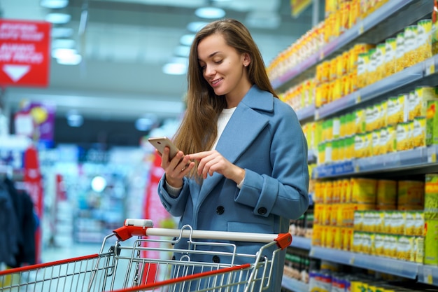 Ritratto di un giovane compratore femminile attraente felice sorridente con il carrello nella navata laterale del negozio con la lista della drogheria sullo smartphone durante l'alimento di acquisto