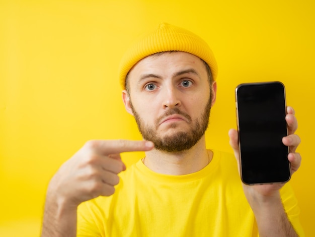 Ritratto di un giovane eccitato, che indica un telefono cellulare con il dito indice. Isolato su sfondo giallo