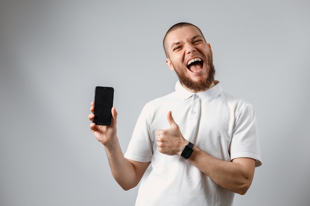 Ritratto di un giovane felice in una maglietta bianca che mostra uno schermo del telefono su gray.