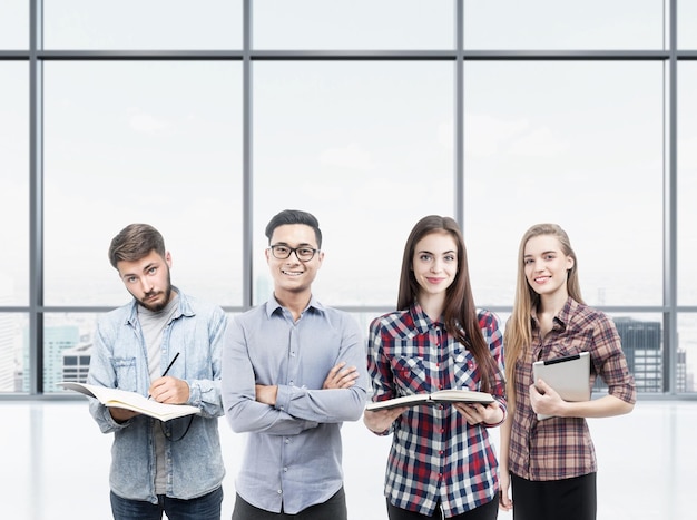 Ritratto di un giovane gruppo di affari che sta con i libri e un computer della compressa in un ufficio vuoto con una finestra panoramica. Concetto di avvio e formazione aziendale. Modello