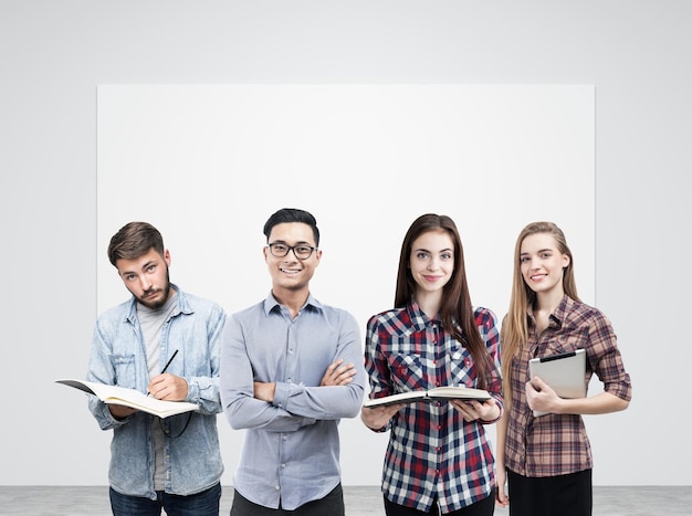 Ritratto di un giovane gruppo di affari che sta con i libri e un computer della compressa vicino a un manifesto in bianco che appende su un muro di cemento. Modello