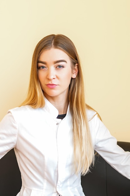 Ritratto di un giovane medico caucasico con capelli lunghi biondi che indossa un camice bianco seduto sul divano nero guardando la telecamera