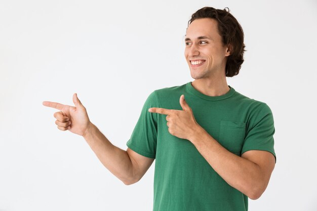 Ritratto di un giovane ottimista che indossa una maglietta di base che ride e punta le dita da parte a copyspace isolato su un muro bianco