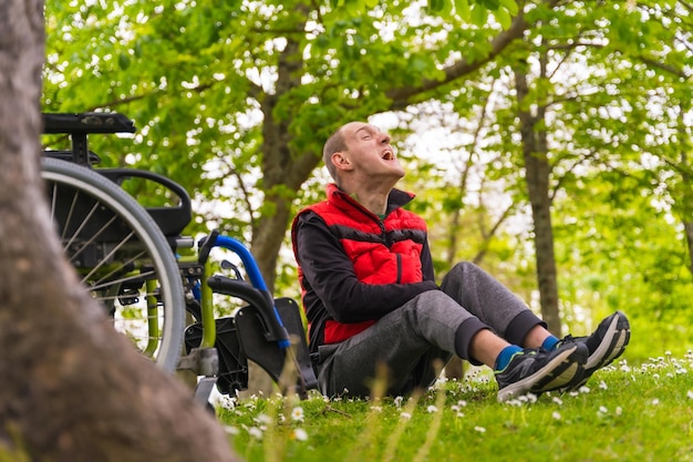 Ritratto di un giovane paralizzato seduto sull'erba accanto a una sedia a rotelle accanto a un albero che ride ad alta voce