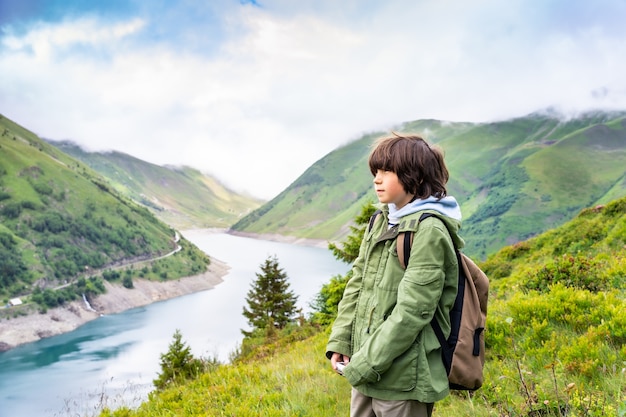 Ritratto di un giovane ragazzo con zaino in una giacca verde in piedi in montagna vicino a un bellissimo lago nelle Alpi francesi, guardando lontano con una faccia seria. Escursionismo con i bambini.