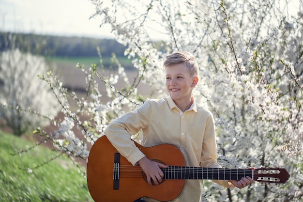 Ritratto di un giovane ragazzo in piedi nel parco di primavera e suonare la chitarra.