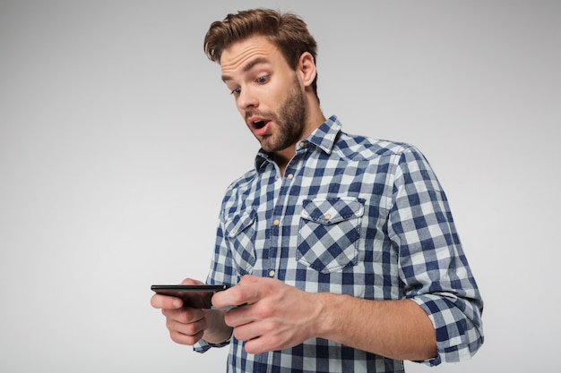 Ritratto di un giovane scioccato che indossa una camicia a quadri che gioca al videogioco sul cellulare isolato su un muro bianco