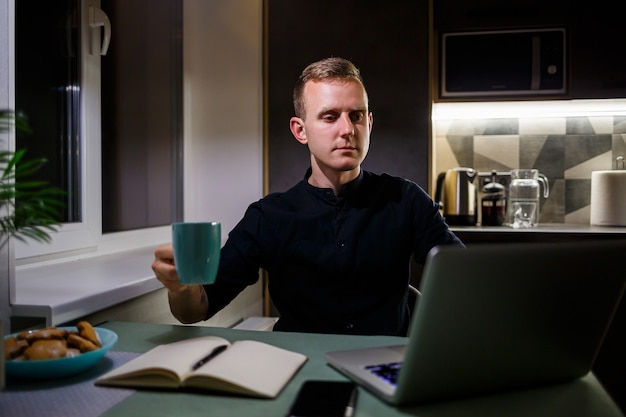 Ritratto di un giovane sorridente che lavora su un laptop da casa e beve caffè da una tazza ed è soddisfatto del suo lavoro. Accogliente appartamento moderno in stile loft