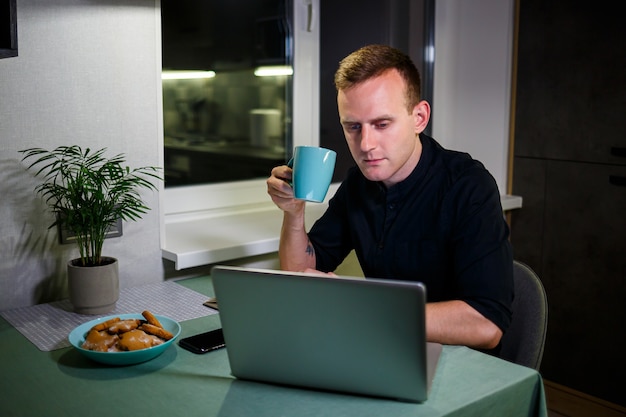 Ritratto di un giovane sorridente che lavora su un laptop da casa e beve caffè da una tazza ed è soddisfatto del suo lavoro. Appartamento moderno e accogliente. Lavoro a casa, concetto di business online