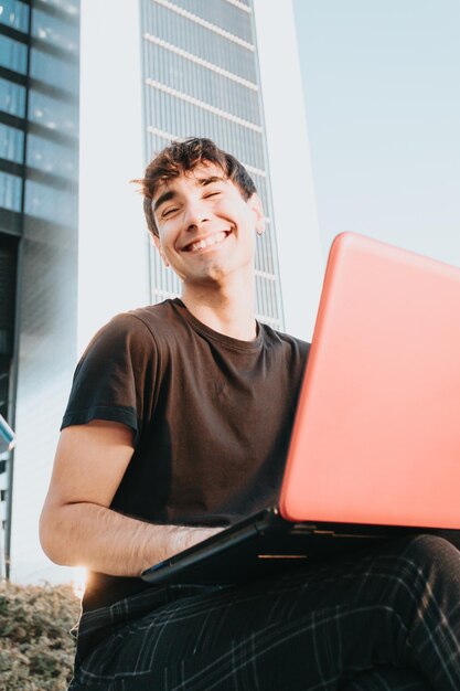 Ritratto di un giovane studente di affari in una camicia nera che sorride alla telecamera mentre si utilizza il laptop facendo una videochiamata in un concetto di business studentesco all'aperto, notebook, edifici aziendali di design moderno