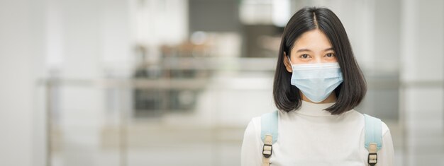 Ritratto di un giovane studente universitario asiatico adolescente in abito casual, tiene in mano un tablet con lo zaino della scuola, indossa una maschera medica e mantiene le distanze sociali durante la pandemia di COVID-19 nel campus universitario