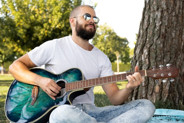 Ritratto di un giovane uomo barbuto e rasato a suonare la chitarra nel parco