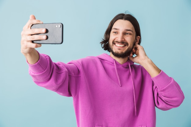 Ritratto di un giovane uomo bruna barbuto felice che indossa una felpa con cappuccio in piedi isolato sul muro blu, facendo un selfie