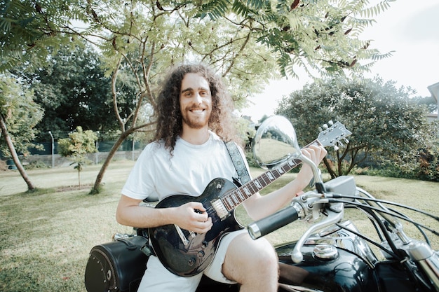 Ritratto di un giovane uomo con i capelli lunghi in una moto che suona la chitarra elettrica mentre si riposa da un viaggio. Concetto di viaggio. Guanti e mani maleducate che tengono una mappa su una moto vecchia scuola.
