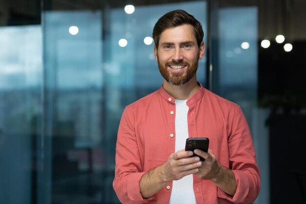Ritratto di un giovane uomo con una camicia rossa in piedi in ufficio con in mano un telefono alla ricerca