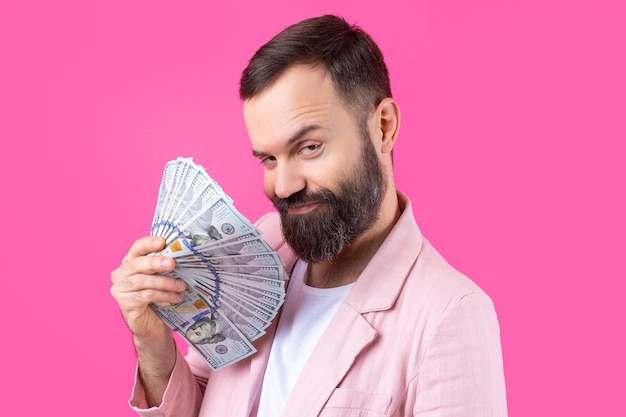 Ritratto di un giovane uomo d'affari soddisfatto con la barba vestito con una giacca rosa che ci mostra le banconote in dollari su uno sfondo rosso dello studio Gusto odore di denaro