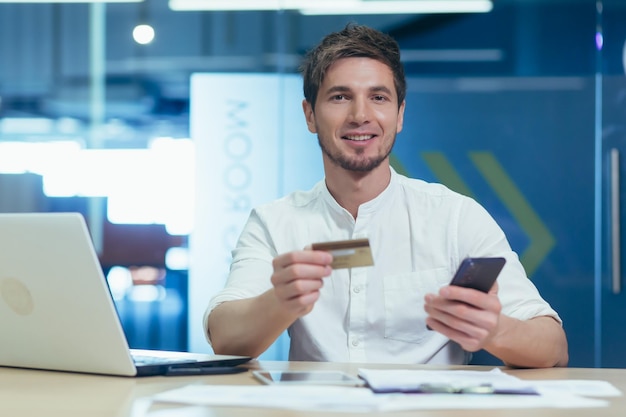 Ritratto di un giovane uomo d'affari sorridente e guardando la telecamera in possesso di un telefono e una carta di credito bancaria per lo shopping in un negozio online